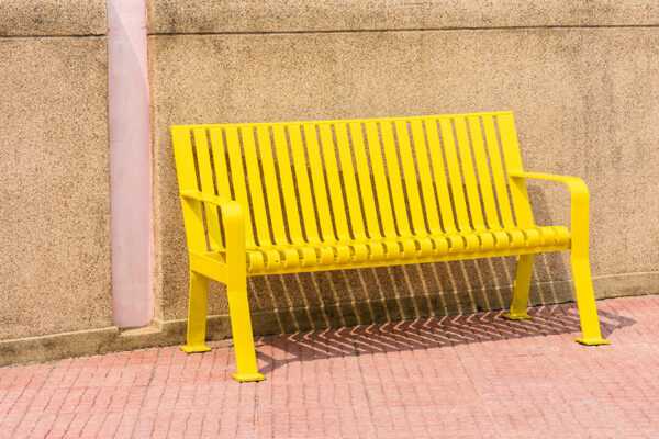 A bright yellow metal bench stands on concrete pavers against a textured beige wall