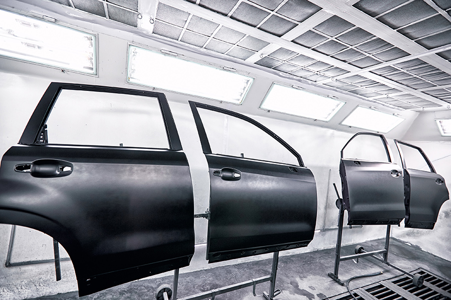 Three black car doors are mounted vertically on stands inside a brightly lit, white automotive paint booth