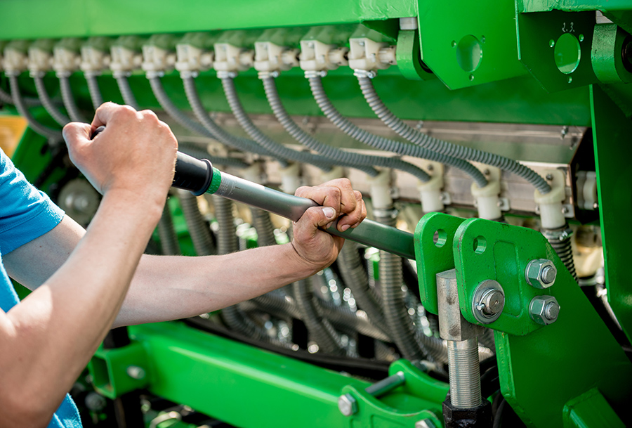 A person uses a wrench to adjust or tighten bolts on a large green agricultural machine with numerous hoses and metal fittings.