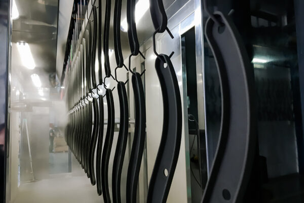 Multiple black metal bars or parts are hanging on hooks in a manufacturing or industrial setting, ready for powder coating