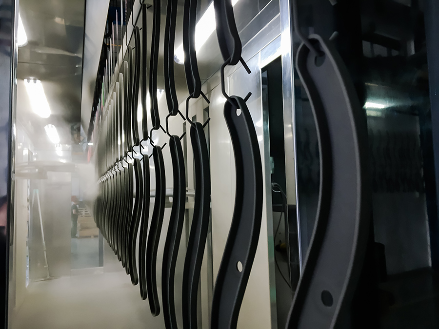 Multiple black metal bars or parts are hanging on hooks in a manufacturing or industrial setting, ready for powder coating
