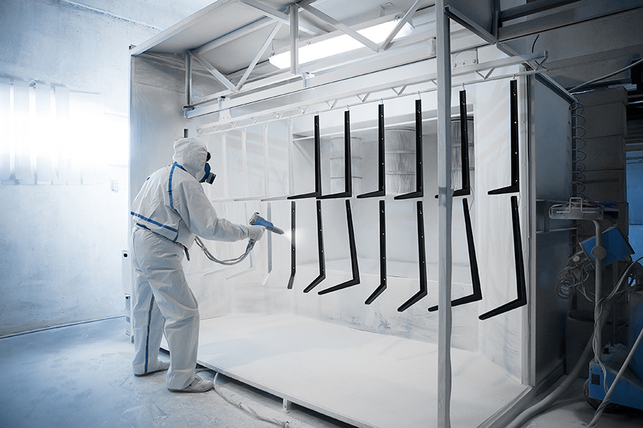 A person in protective gear and a respirator uses a spray gun to powder coat metal parts hanging in a booth