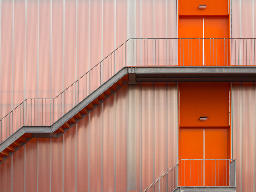 A modern building facade with bright orange doors and matching railings. A metal staircase runs diagonally across the image, contrasting with the translucent, vertically striped wall panels.